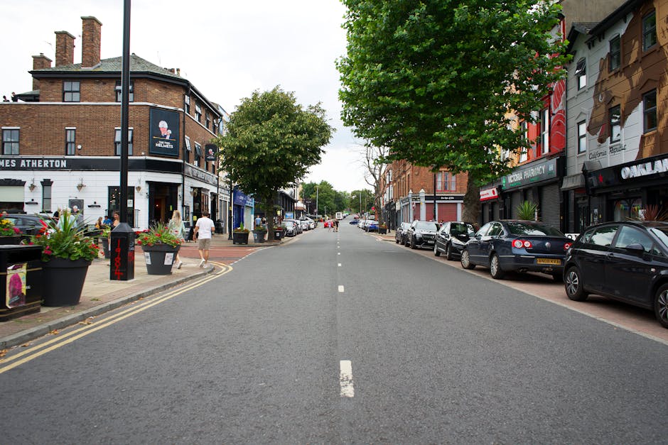 A wide street scene in Marks Gate with parked cars along both sides of the road and commercial buildings, including a pub named 'James Atherton' on the left and various retail outlets on the right. The street is lined with trees and potted plants on the pavements, with pedestrians walking along the sidewalks. The road appears clear of traffic, providing space for loading or unloading during a home relocation or furniture transport. This setting reflects an urban environment suitable for professional removals, such as those offered by Man with Van Marks Gate, involving loading and packing operations for house moves or business relocations. The scene is well-lit with natural daylight, emphasizing the calm, organized street environment necessary for efficient moving services.