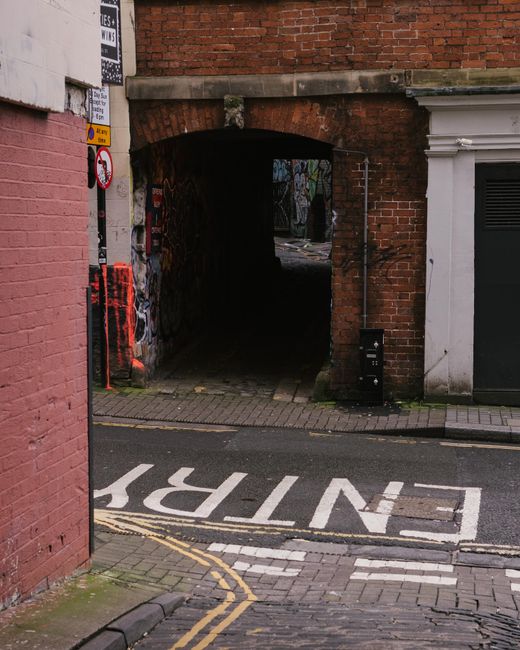 Photograph of a narrow brick archway leading from a busy street into an alleyway behind buildings, with a paved footpath and a marked 'ENTRY' area on the road in front. To the left of the arch, there is a pink-painted brick wall with traffic and parking signs attached. Inside the alleyway, visible through the arch, are graffiti-covered walls and a dark, shadowed passage. The street in the foreground has a curb, tactile paving, and a parking restriction zone with dashed yellow lines. To the right of the arch, part of a white building with decorative moldings and an air vent is visible. The scene suggests a location suitable for home relocation or furniture transport, with possible use by a professional removals service like Man with Van Marks Gate for moving logistics and loading processes.