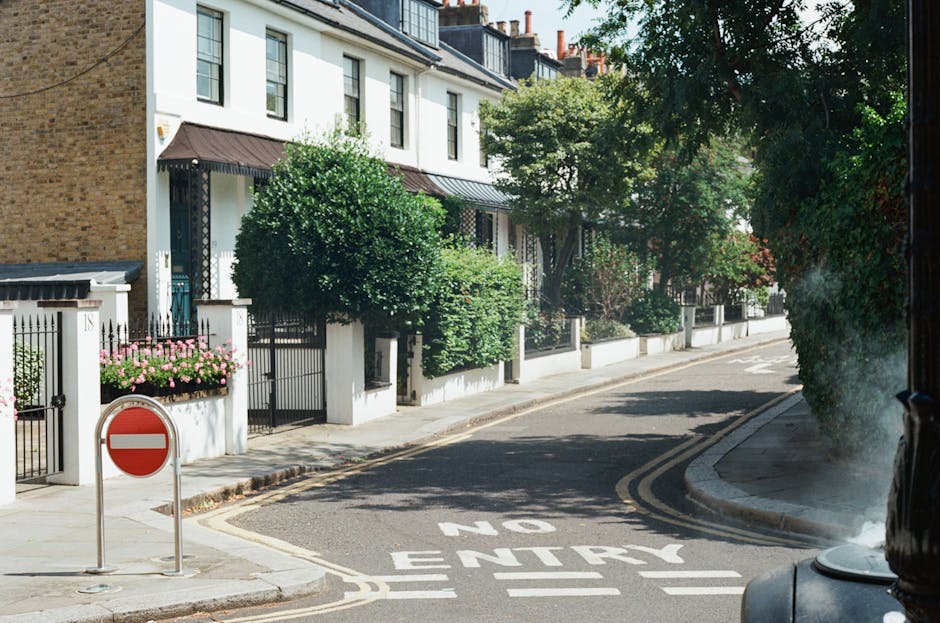 A wide street scene in Marks Gate with parked cars along both sides of the road and commercial buildings, including a pub named 'James Atherton' on the left and various retail outlets on the right. The street is lined with trees and potted plants on the pavements, with pedestrians walking along the sidewalks. The road appears clear of traffic, providing space for loading or unloading during a home relocation or furniture transport. This setting reflects an urban environment suitable for professional removals, such as those offered by Man with Van Marks Gate, involving loading and packing operations for house moves or business relocations. The scene is well-lit with natural daylight, emphasizing the calm, organized street environment necessary for efficient moving services.