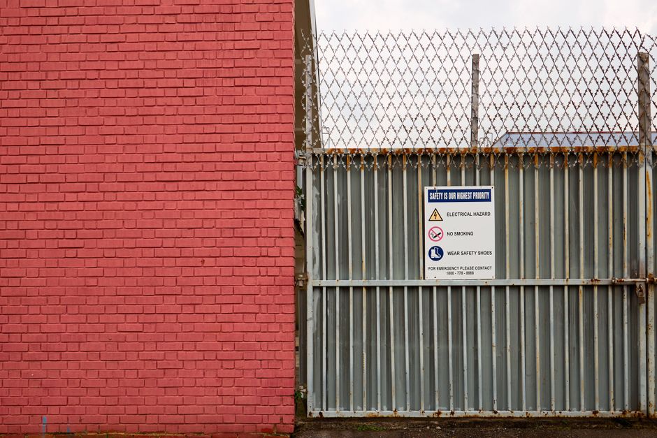 A close-up view of a red brick wall on the left side and a large metal sliding gate on the right side, which is part of a property boundary. The metal gate is made of vertical corrugated steel panels with signs of rust and corrosion, topped with barbed wire for security. Mounted on the gate is a white sign displaying safety instructions, including warnings about electrical hazards, no smoking, and the requirement to wear safety shoes, along with a contact number for emergencies. Behind the gate, there is a white panel, possibly covering windows or access points, and the gate is situated on a paved pavement area. This scene is inside a commercial or industrial premises, and the surroundings suggest an area often used for loading and unloading during house removals or furniture transport. The setting reflects typical logistics involved in home relocation or moving services, where secure fencing and guidance signage prevent hazards during loading activities associated with house removals by companies such as Man with Van Marks Gate.
