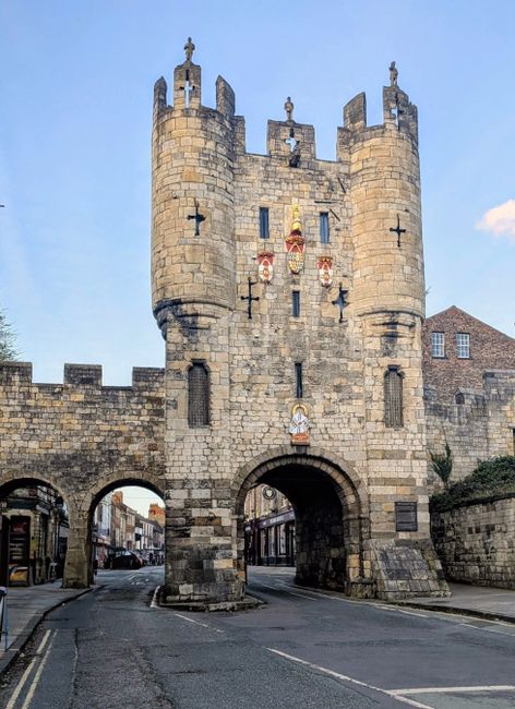 A historic stone gatehouse with a central archway serving as an entrance to a town or city. The structure features crenellated battlements at the top, decorative shields and heraldic symbols on the fa