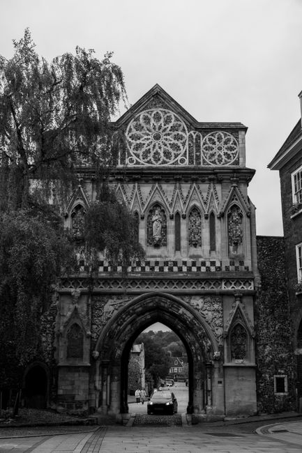 A historic stone gatehouse with a central archway serving as an entrance to a town or city. The structure features crenellated battlements at the top, decorative shields and heraldic symbols on the fa