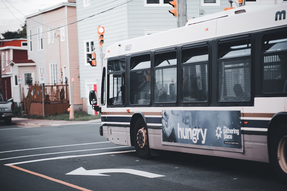 A white public transit bus parked at a curb on a residential street during daylight hours, with its doors closed and windows visible. The bus features black-framed windows along the side and an advertisement on the lower section displaying the word 'hungry' and the logo for Gathering Space. The bus is situated near a pedestrian crossing with white painted lines and a directional arrow on the road. Surrounding the bus are multi-storey houses painted in pastel shades of pink, grey, and white, with some houses having wooden fences, small gardens, and front lawns. There are streetlights, traffic signals, and overhead wires visible above the street. In the background, a black car is parked along the side of the road, and the scene suggests urban or suburban surroundings. The image captures a moment during a home relocation or furniture transport process managed by Man with Van Marks Gate, with a focus on the vehicle as part of the moving logistics.