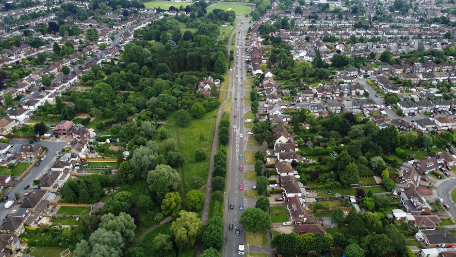 A quiet residential street with a narrow, straight tarmac road marked with a central white dashed line, flanked by residential houses on the left and a fields on the right. Tall, leafless trees cast shadows across the road, indicating early spring or late winter. The pavement on the left side is partially excavated, possibly for utility work or preparation for loading activities. A brick wall with a small gate marks the entrance to a property on the left, while the right side features a grassy verge with bushes and a wooden fence. The scene is well-lit with clear blue sky overhead, and no vehicles or people are visible, suggesting a calm, low-traffic environment suitable for local house removals or furniture transport. Occasionally, Man with Van Marks Gate arranges home relocation services, including loading and packing, in settings like this during their moving operations. 