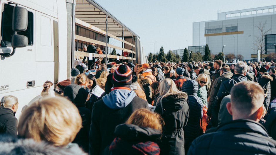 A large group of people gathered outside in an urban area during daytime, standing on a pavement near a white moving truck with an open side showing wooden pallets and packing materials, including cardboard boxes and blankets, inside. The scene includes diverse individuals wearing jackets, coats, and hats, some carrying or handling boxes and bags. The background features modern office buildings with large windows and trees, under a clear sky. The image captures the busy environment typical of busy moving day logistics, illustrating the importance of efficient furniture transport and packing during home relocation. Man with Van Marks Gate services are associated with handling such movements smoothly, often involving loading processes and managing delays during house removals.
