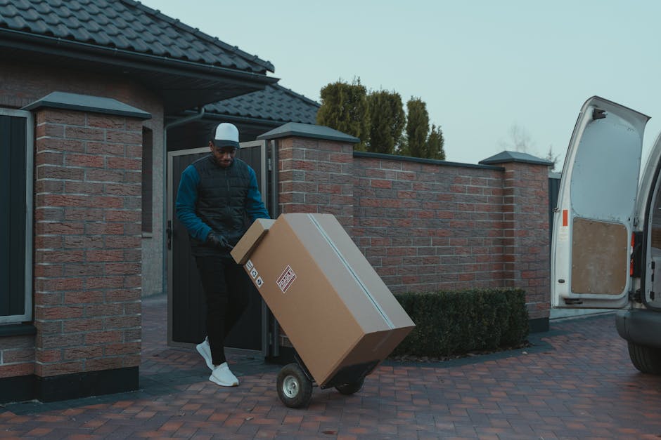 A man wearing a blue and black jacket, dark trousers, and a white cap is loading a large cardboard box secured with packing tape onto a hand truck or dolly. He is positioned at the entrance of a residential property, with a black gate and brick pillars behind him. The house has a dark tiled roof, and there are tall, green trees visible in the background. To the right, an open white van with a partially visible interior is parked on a brick driveway, ready for furniture transport or packing and moving activities. The scene is outdoors in natural daylight, capturing a home relocation process involving furniture transport, with various cardboard boxes and packing materials used during the loading process. This image illustrates a step in the logistical aspect of house removals, supported by the services of Man with Van Marks Gate, focused on efficient moving and logistical planning.