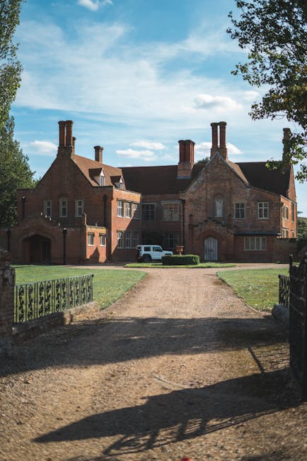 A large red-brick estate with multiple chimneys and sash windows is visible in the background under a partly cloudy sky. In the foreground, a gravel driveway leads up to the building, bordered by black metal fencing and a patch of green grass. A white van is parked near an entrance on the property's front side. The scene is illuminated by natural daylight with some trees visible on the right side, providing partial shade. The image captures a typical scene associated with home relocation or furniture transport, as part of a house removals process managed by Man with Van Marks Gate, showing the exterior environment where furniture and boxes might be loaded or unloaded during a move.