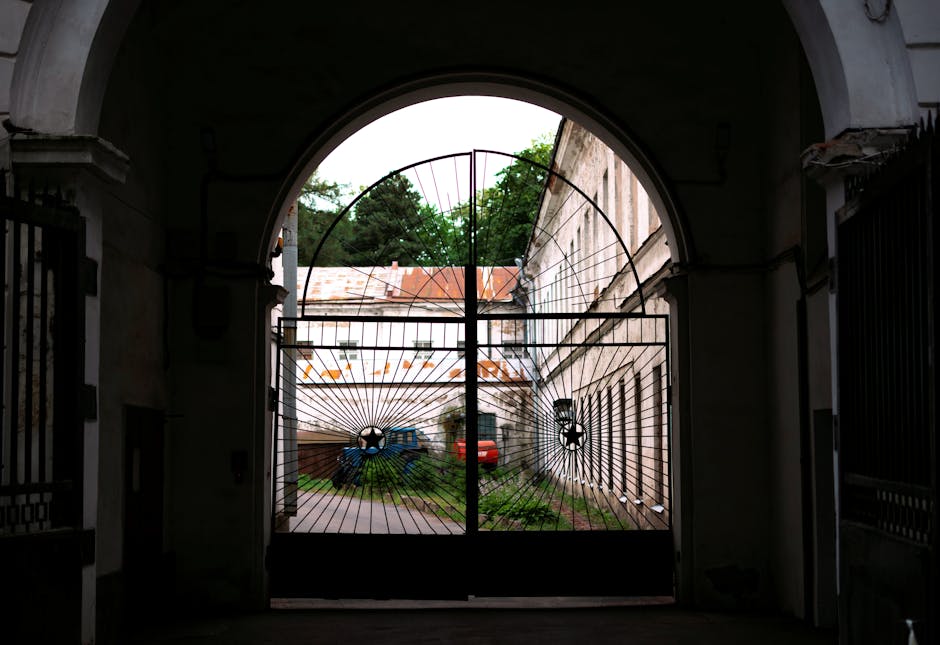 A black metal gate with vertical bars and decorative scrollwork at the bottom, partially obscuring a view of a residential area through the bars. Attached to the gate is a small sign reading 'PRIVATE,' indicating restricted access. The gate is positioned at the entrance of a property, with a pathway visible beyond, leading to houses with multiple windows and surrounding greenery. The scene is outdoors, captured during daytime with natural light reflecting off the metal surface. This image relates to house removals and moving services, emphasizing secure property access in the context of local home relocations, such as those undertaken by Man with Van Marks Gate during furniture transport or packing and moving processes.