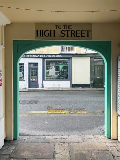 View through a green-painted archway on a covered passage leading from an interior space to the high street, with a sign above reading 'TO THE HIGH STREET.' Outside, the paved sidewalk runs parallel t