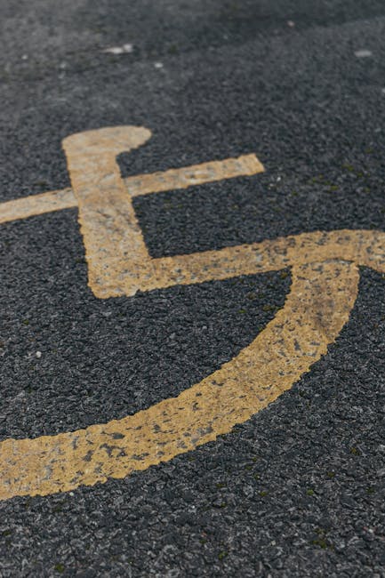 A close-up image of a yellow wheelchair symbol painted on a dark asphalt surface, indicating a designated accessible parking space. The paint appears slightly worn with visible texture and small cracks in the material. In the background, a section of the parking lot pavement is visible with small gravel and rough surface details. This image was likely taken outdoors, near a building entrance or driveway used for home relocation or moving purposes. It supports content related to house removals and moving logistics by illustrating accessible parking for vans or vehicles involved in furniture transport, packing, or loading processes. Man with Van Marks Gate may use such designated spaces when providing removals services, ensuring efficient access for loading and unloading furniture and boxes during the home relocation process.