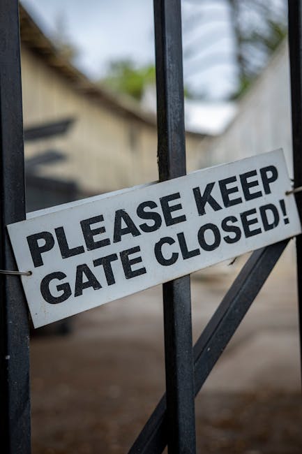 Close-up of a black metal gate during a home relocation, with a white sign attached that reads 'PLEASE KEEP GATE CLOSED!'. The gate appears sturdy and is part of a driveway or entrance area, with a gravel or paved surface visible beneath. In the background, blurred elements suggest a residential setting, possibly a house or outbuilding, with some greenery and trees indicating an outdoor environment. The sign indicates the importance of keeping the gate secured during the moving process, which is relevant to furniture transport and packing logistics for house removals. The setting is well-lit with natural daylight, emphasizing that the gate is part of a property access point involved in house removal activities, managed by Man with Van Marks Gate, as referenced on their website's page about local moving tips.