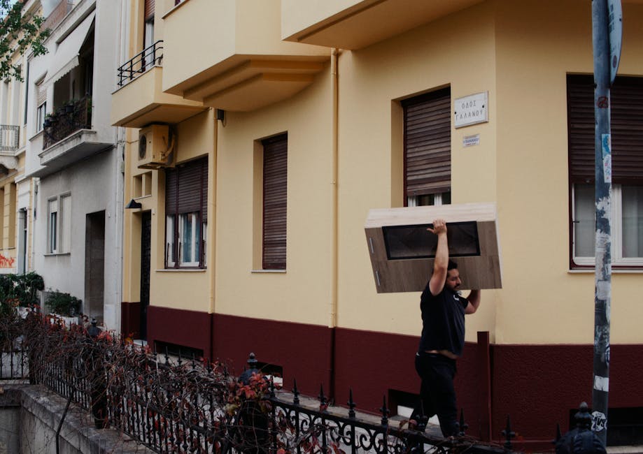 A man with a beard dressed in a dark uniform is carrying a large wooden cupboard with a black glass window through a narrow doorway on the front of a pale yellow residential building in Marks Gate. The building has three windows with closed brown shutters, some with flower boxes on the metal balconies, and an address plaque. He is walking on a sidewalk bordered by a decorative wrought iron fence, and the scene is illuminated by natural daylight. The sidewalk and building's entrance are part of a home relocation or furniture transport process coordinated by Man with Van Marks Gate, with the man preparing to load or unload the bulky item for a house move, demonstrating the logistical aspects of packing and moving within flats that feature narrow lifts and limited access areas.