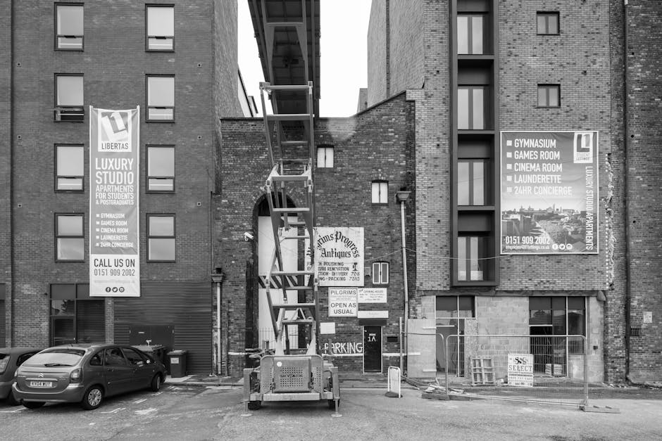 A black and white photograph capturing the loading process of home relocation or furniture transport outside a multi-storey residential building in Marks Gate. The image shows a large, mobile scaffold tower positioned on a paved area directly in front of the building's entrance, with its platform extended near a narrow lift shaft. The building is constructed with brickwork and features multiple small windows, some of which have window air conditioning units. Two large vertical banners advertising local amenities such as a gymnasium, cinema room, and laundrette are mounted on the building's wall. In the foreground, several cars are parked, and a few signs indicating parking restrictions and building services are visible near the entrance. The scene includes elements typical of moving logistics, such as protective blankets and items being carried or prepared for loading, with a focus on the careful handling of household belongings during the moving process. Man with Van Marks Gate is visible as the service provider associated with house removals and associated logistics.
