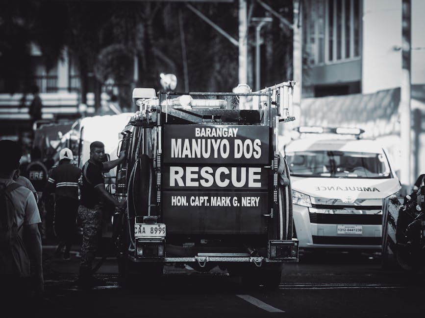 A black-and-white photograph depicting a rescue vehicle parked on a city street during daytime, surrounded by emergency personnel including firefighters and medical staff. The rescue truck, prominentl