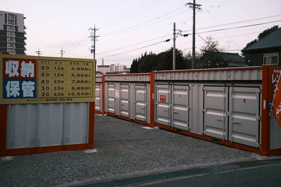 Photograph of a storage facility consisting of multiple grey metal lockers secured with padlocks, situated outdoors on a gravel surface next to a sign displaying pricing for various storage unit sizes in Japanese yen. The sign is mounted on a white metal panel with orange borders, indicating storage options such as 6D, 5D, 4D, 3D, and 2D, with respective prices and usage details. In the background, residential buildings, utility poles, and overhead power lines are visible under a clear sky, suggesting an urban or suburban environment. The setup appears to be part of a logistics route for home relocation or furniture transport, consistent with the services offered by Man with Van Marks Gate, who handle removals and packing. The scene captures the process of storing belongings temporarily during house moves, with the lockers ready for securing packed boxes or furniture packaging materials, reflecting typical moving logistics involving loading or unloading activities.